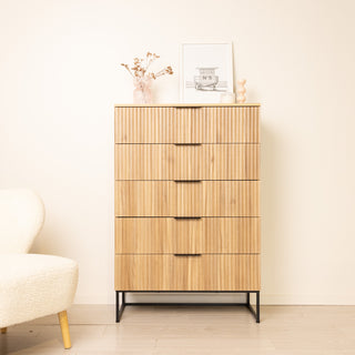 Lifestyle view of the Reede Tallboy 5-Drawer in oak finish, featuring vertical fluted drawer fronts, black handles, and slim black legs. Styled in a modern living room with a cream chair, framed artwork, and decor pieces on top.