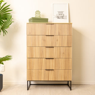 Front view of the Reede Tallboy 5-Drawer dresser in oak finish, featuring vertical fluted drawer fronts, slim black handles, and elevated black metal legs, styled with decor accents in a modern living space.
