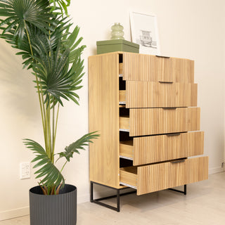 Reede Tallboy 5-Drawer dresser, 80 × 39 × 120 cm, in warm oak finish with vertical fluted drawer fronts and slim black metal base, shown with drawers partially open beside a tall potted plant.