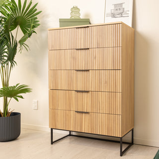 Side angle view of the Reede Tallboy 5-Drawer dresser in oak finish, showcasing vertical fluted drawer design, slim black handles, and raised black metal legs, styled in a modern home with indoor plants and wall art.