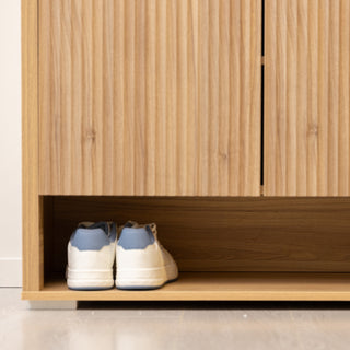 Renne shoe cabinet in oak finish showing the open bottom shelf with a pair of sneakers neatly stored. The design combines vertical fluted doors with a practical base compartment, providing extra space for everyday shoes while keeping the entryway tidy and stylish.
