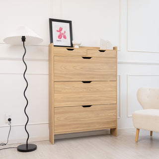 Risa slim shoe cabinet in oak finish styled in a modern living space with a floor lamp and armchair, featuring three tilt-down compartments and two top drawers for hallway storage.