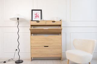 Risa Slim Shoe Cabinet in oak finish showing tilt-down compartment open, slim tall design placed in a bright room with white chair and floor lamp.