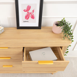 Close-up of the Ruelle 6 Drawer dresser in oak finish, showing a partially open drawer with a Dior book inside. The dresser features brass gold handles, a fluted side design, and is styled with a framed artwork and a pink planter on top.