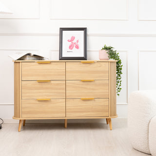 Front view of the Ruelle 6 Drawer dresser in oak finish, featuring brass gold handles and rounded fluted sides. Styled with a framed artwork, decorative tray, and potted plant on top, placed in a bright modern room with a white armchair beside it.