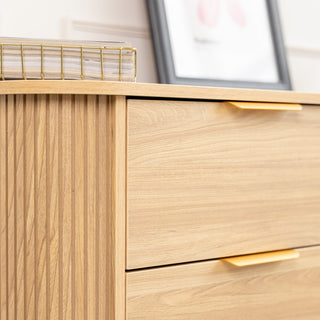 Close-up side detail of the Ruelle 6 Drawer dresser in oak finish, highlighting the vertical fluted design, brass gold handles, and smooth drawer surface. A decorative tray and framed artwork sit on top, adding a modern touch.