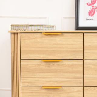 Close-up of the Ruelle 6 Drawer dresser in oak finish, showing the brass gold handles on the top left drawers and the fluted side panel. A gold wire tray with books and a framed artwork sit on the dresser’s surface, styled in a modern interior.