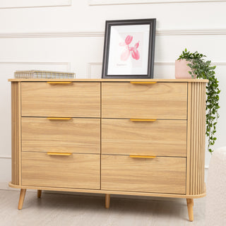 Front view of the Ruelle 6 Drawer dresser in a warm oak finish, featuring sleek brass gold handles and vertical fluted side panels. The dresser sits on tapered wooden legs, styled with a framed artwork and a plant on top.