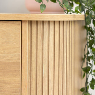 Close-up of the Ruelle 6 Drawer dresser’s vertical fluted side panel in a warm oak finish, highlighting the fine craftsmanship and smooth rounded edge with trailing green plant decor on top.