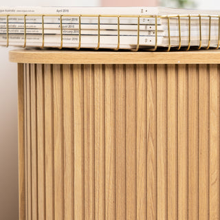 Close-up of the Ruelle 6 Drawer dresser showing the rounded oak top edge and vertical fluted side panel, styled with a gold wire tray holding stacked magazines for a modern touch.