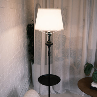 Slate Tray Floor Lamp featuring a soft fabric shade, decorative black metal stem and round tray shelf, placed beside sheer curtains and a sofa in a calm living room setting.