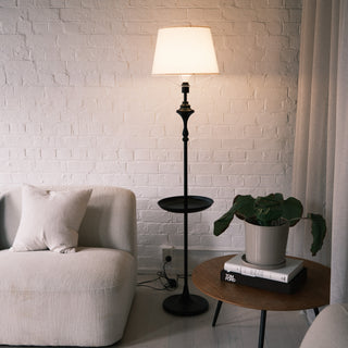 Slate Tray Floor Lamp with a warm fabric shade and matte black tray table, placed beside a cream sofa and styled with a wooden side table and indoor plant against a white brick wall.