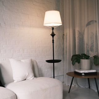 Slate Tray Floor Lamp positioned beside a soft cream sofa, featuring a warm fabric shade and matte black tray table, styled in a calm living room with white brick walls and neutral decor.