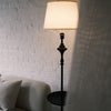 Slate Tray Floor Lamp positioned beside a textured cream sofa, showing its warm fabric shade and matte black sculpted stem. The built in tray table is partly visible, with soft natural light filtering through curtains against a white brick wall.
