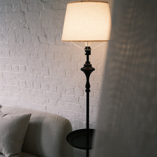Slate Tray Floor Lamp positioned beside a textured cream sofa, showing its warm fabric shade and matte black sculpted stem. The built in tray table is partly visible, with soft natural light filtering through curtains against a white brick wall.