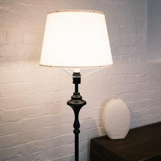 Close up of the Slate Tray Floor Lamp showing the warm fabric shade and matte black sculpted stem. The lamp sits beside a wooden console and a white ceramic vase against a textured white brick wall, highlighting its classic detailing and soft ambient lighting.