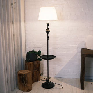 Slate Tray Floor Lamp featuring a matte black metal body, round tray shelf, and warm fabric shade. The lamp is shown in a cosy living room corner with natural light, wooden stools, indoor plant, and a textured white brick wall, highlighting its functional tray design and soft ambient lighting.