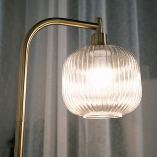Close up of the Solara Fluted Lantern Floor Lamp, showing the softly glowing fluted glass shade and brushed brass arm against sheer curtains.