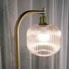 Close up of the Solara Fluted Lantern Floor Lamp showcasing its softly curved glass shade, warm illuminated glow and brushed gold arm, with sheer curtains in the background adding a gentle, elegant atmosphere.