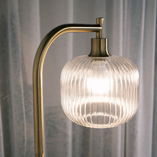 Close up of the Solara Fluted Lantern Floor Lamp showcasing its softly curved glass shade, warm illuminated glow and brushed gold arm, with sheer curtains in the background adding a gentle, elegant atmosphere.