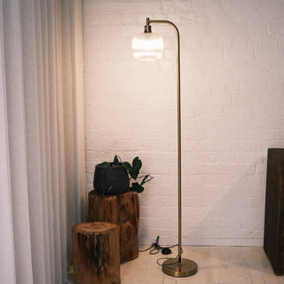 Solara Fluted Lantern Floor Lamp placed in a modern living space, featuring a tall brushed brass frame, rounded fluted glass shade, and warm ambient lighting beside natural wood stools and soft curtains.