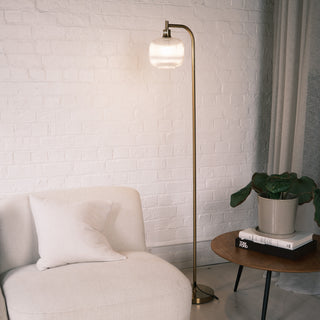 Solara Fluted Lantern Floor Lamp standing beside a white curved sofa, casting a soft warm glow against a textured brick wall, with a wooden side table, stacked books, and a potted plant completing the cosy lounge scene.