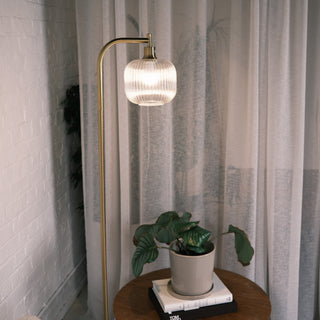 Solara Fluted Lantern Floor Lamp positioned beside soft sheer curtains, featuring a warm brass stand and ribbed glass shade, softly illuminating a round wooden side table with a potted plant and stacked books.