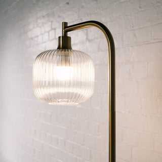 Close up of the Solara Fluted Lantern Floor Lamp highlighting its sculpted fluted glass shade and brushed brass arm, softly illuminated against a white brick wall for a warm and elegant look.