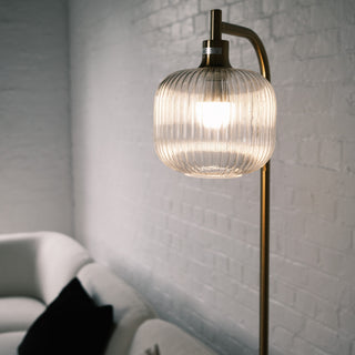 Illuminated fluted glass shade of the Solara Lantern Floor Lamp, showcasing its rounded sculptural form and brushed brass frame against a soft white brick interior setting.
