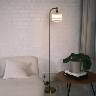 Solara Fluted Lantern Floor Lamp featuring a brushed brass metal frame and sculpted fluted glass shade, placed beside a soft cream sofa in a modern living room with white brick walls and a wooden side table. The lamp emits a warm ambient glow, highlighting its curved silhouette and minimalist design.