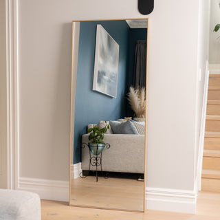 The Solene X full-length mirror, featuring a 170 x 70 cm wooden PS frame in a natural oak finish, placed against a hallway wall beside a staircase. Its double silver glass surface reflects a cosy lounge with blue walls, artwork, and soft decor, showing how the mirror can open up and brighten smaller living spaces.