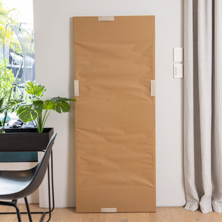 The back of the Solene X full-length mirror (170×70 cm) wrapped in protective brown paper, showing its secure mounting design that supports both vertical and horizontal hanging. Photographed beside a window with indoor plants and soft curtains, this image highlights the mirror’s versatile installation and careful packaging.
