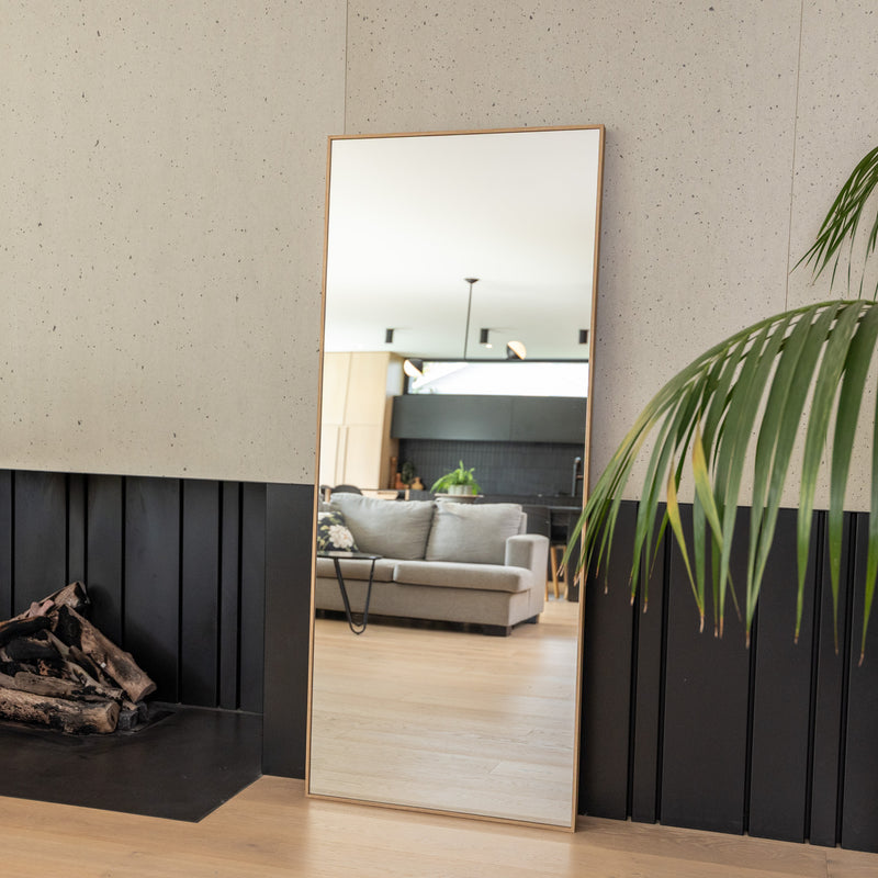 The Solene X Straight-Edged Full-Length Mirror (170×70 cm) with a 45 mm wood-look PS frame in a warm oak tone. Featuring 4 mm double silver mirror glass for exceptional clarity, this modern rectangular mirror leans gracefully against a textured wall in a contemporary living room with a sofa, fireplace, and green plants, reflecting a bright open space bathed in natural light.