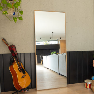 The Solene X full-length mirror (170×70 cm) featuring a 45 mm wooden-look PS frame with 4 mm double silver glass. Placed beside a guitar and indoor plants in a contemporary living room, the mirror reflects a bright open interior with a neutral sofa and black accent wall, highlighting its sleek rectangular shape and natural wood tone.