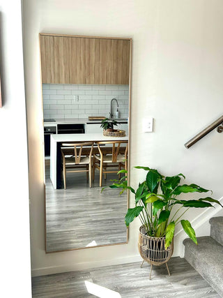 Customer photo of the Solene X full-length mirror with a wooden PS frame and 4mm double silver glass, styled in a bright, modern home interior. The mirror reflects a warm kitchen and dining space with light timber accents, creating an elegant and natural atmosphere complemented by a lush indoor plant.
