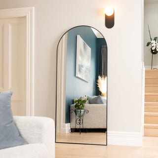 The Titan X arched full-length mirror leaning gracefully against a modern living room wall, reflecting a cosy interior with blue feature wall, artwork, and a soft grey sofa. Its 32 mm aluminium frame adds a sleek, durable touch that enhances the room’s spacious and refined aesthetic.