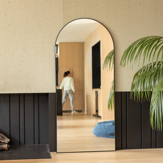 The Titan X arched full-length mirror placed in a contemporary hallway, reflecting a warm timber interior and a person walking in the background. The matte black 32 mm aluminium frame and clear reflection highlight the mirror’s refined design, blending durability with timeless elegance.