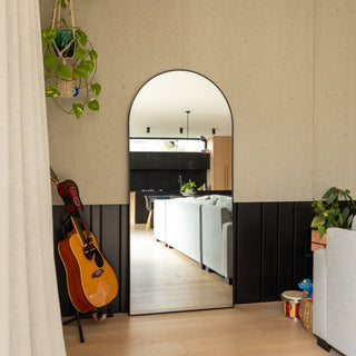 Titan X arched full-length mirror leaning against a textured wall, reflecting a cosy modern living room with a grey sofa and wooden interior. A guitar, hanging plants, and a woven basket nearby create a relaxed, homely atmosphere, highlighting the mirror’s elegant curve and sleek black aluminium frame.