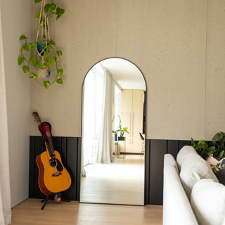 The Titan X arched full-length mirror placed against a modern textured wall, reflecting a bright hallway filled with natural light. A hanging plant and acoustic guitar beside the mirror add warmth and personality, while the slim black 32 mm aluminium frame enhances the sleek, contemporary aesthetic.