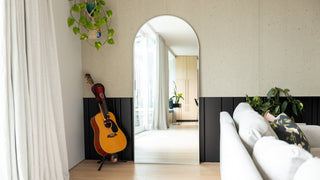 Titan Alba Arched Full-Length Mirror placed in a modern living room with a light oak floor, grey sofa, and acoustic guitar beside it. The mirror’s elegant 30mm slim black frame and ultra-clear double silver 4mm glass reflect the bright interior and natural light, highlighting its clean arch design and contemporary appeal.
