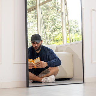 The Titan X arched full-length mirror captures a clear, true-to-life reflection as a man reads comfortably in a bright living room. Its 4mm distortion-free glass and sleek aluminium frame highlight modern craftsmanship and offer a calm, reflective moment in any New Zealand home interior.