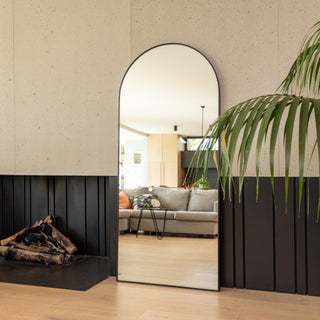 The Titan X arched full-length mirror positioned beside a fireplace, reflecting a stylish living room with a grey sofa, soft cushions, and warm wooden flooring. Its slim 32 mm black aluminium frame and seamless arch design add a refined, contemporary touch to the bright and inviting space.