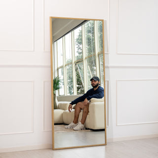 The Tresson 180x80cm gold full-length mirror with a detailed braided PS frame and clear 4mm glass, leaning against a white wall in a bright room. The reflection captures a relaxed man seated on a cream sofa beside large windows and lush greenery, highlighting the mirror’s elegant design and natural lighting effect for modern New Zealand homes.