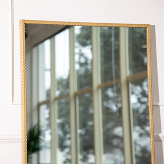 Close-up view of the Tresson gold full-length mirror frame showing its elegant PS material with a textured braided pattern. The 4mm clear glass reflects soft natural light and greenery through tall windows, highlighting the mirror’s refined craftsmanship and luxurious gold finish ideal for modern New Zealand homes.