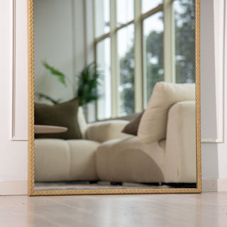 Bottom view of the Tresson gold full-length mirror showing its elegant braided PS frame resting neatly against a light wooden floor. The mirror reflects a beige sofa and greenery near a bright window, highlighting its 4mm clear glass and timeless design perfect for modern New Zealand living spaces.