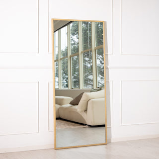 Tresson 180x80cm full-length mirror featuring a gold PS frame with elegant straight edges, reflecting a bright modern living space with large windows and a cream sofa. Designed for wall mounting or floor display, this 4mm clear glass mirror adds a touch of luxury and spaciousness to New Zealand homes.