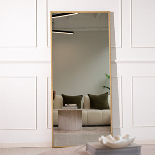 Tresson 180x80cm gold full-length mirror with a refined straight-edged PS frame and 4mm clear glass, leaning gracefully against a white panelled wall. The reflection captures a cosy lounge with green cushions and soft lighting, adding a touch of luxury and warmth to any New Zealand home interior.