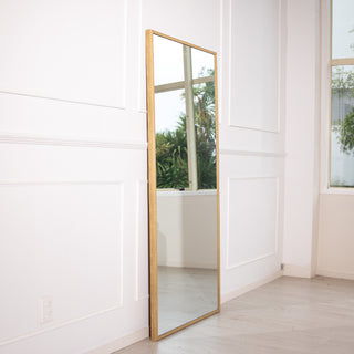 Side view of the Tresson full-length mirror 180x80cm featuring a slim gold PS frame and clear 4mm glass. The mirror leans elegantly against a white wall near large windows, reflecting greenery outside — perfect for adding brightness, depth, and timeless charm to New Zealand home interiors.