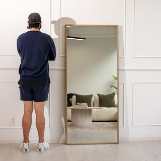 A modern gold full-length mirror from C&F Creation, the Tresson 180x80cm features a sleek PS frame with braided detailing and 4mm clear glass. The mirror is shown leaning against a white wall beside a person, reflecting a cosy beige sofa and green cushions — ideal for creating depth and warmth in contemporary New Zealand interiors.