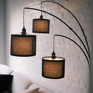 Trilogy 3-Light Floor Lamp featuring three staggered drum shades. The black woven fabric texture is clearly visible. Elegant curved arms extend from a sturdy central pole. Functional ambient lighting for reading or relaxing.
